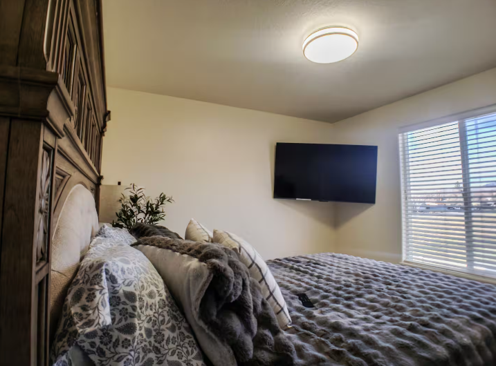 Bedroom with ornate dark wood carved headboard, wall-mounted TV, faux fur throw and natural light from window