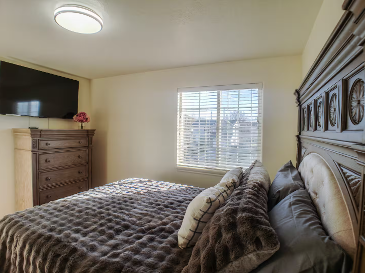 Bedroom with ornate dark wood carved headboard, wall-mounted TV, tall dresser and natural light from window