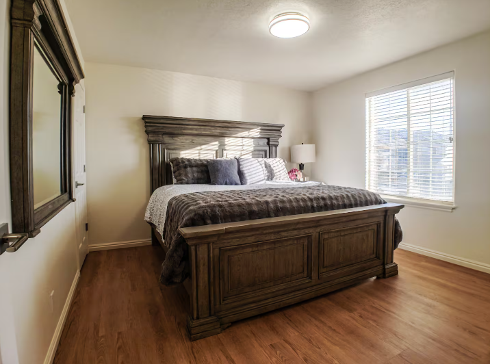 Spacious bedroom with large ornate dark wood bed frame, framed mirror, warm lamp and hardwood floors