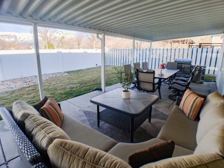 Large covered patio with wicker sectional, dining table for 8, BBQ grill and Wasatch Mountain views beyond white vinyl fence