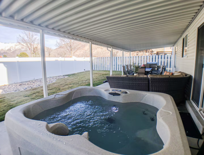 Private hot tub under covered patio with wicker sectional seating, white vinyl fence and Wasatch Mountain views