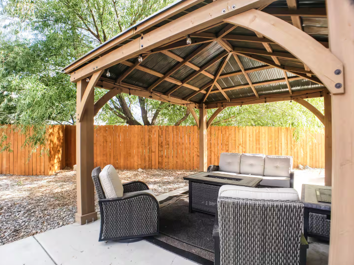 Cedar gazebo exterior with metal roof, string lights, wicker lounge chairs and fire pit table in fenced backyard