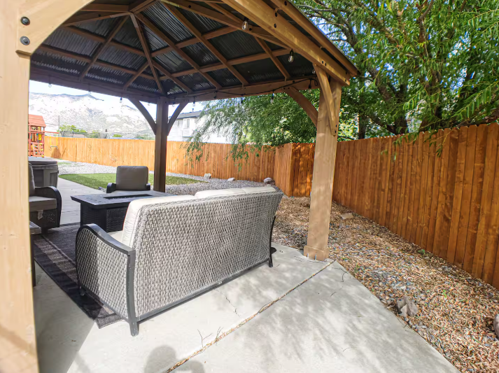 Covered cedar gazebo with wicker sectional sofa, fire pit table and mountain views through the fenced backyard