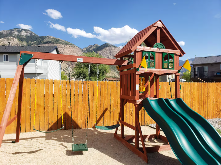 Large wooden kids playground with swing set, green slide, playhouse and mountain backdrop in fenced backyard