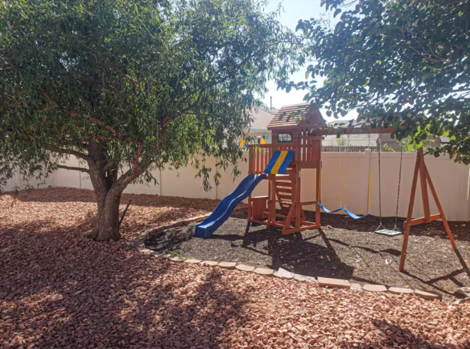 Fenced backyard with wooden playground set, slide, swings and mature shade tree