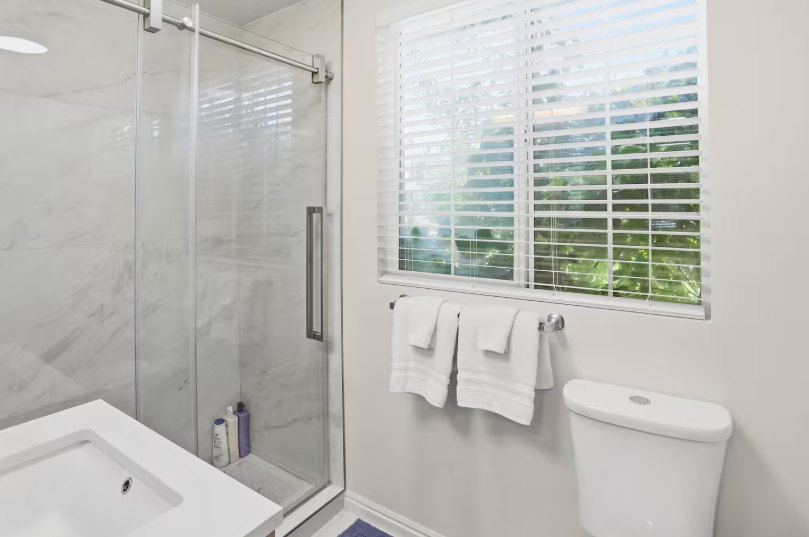 Bright bathroom with marble tile walk-in shower, glass sliding door, white sink vanity, towel bar and window with garden view