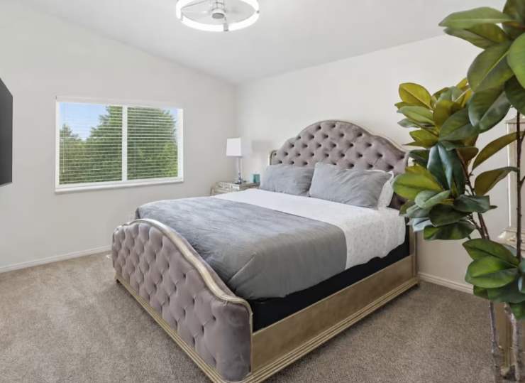 King bedroom with tufted velvet headboard, gray bedding, wall-mounted TV, nightstand lamp and large window with garden view