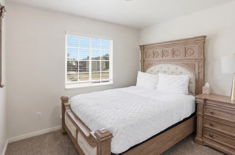 Queen bedroom with ornate carved wood bed frame, white quilted bedding, wood dresser and bright window with plantation shutters