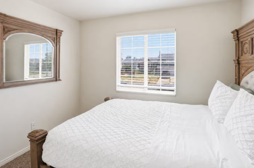 Queen bedroom with ornate carved wood bed frame, white quilted bedding, arched wood mirror and bright window with neighborhood view
