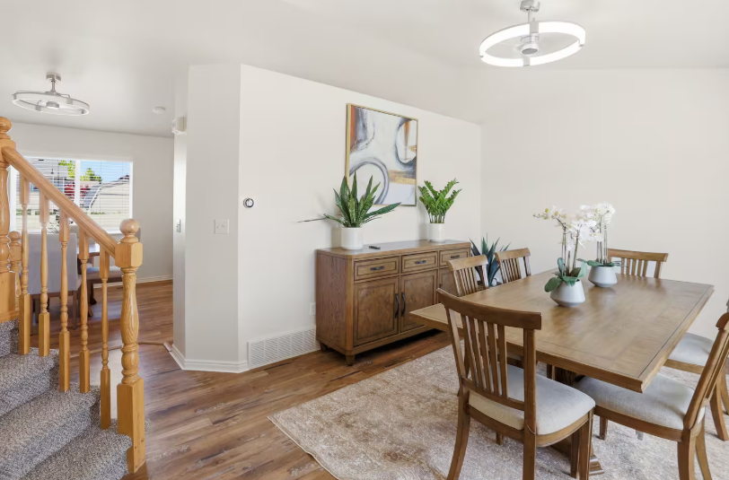 Open dining area with round wood table, four gray upholstered chairs, wood staircase railing and large window with natural light