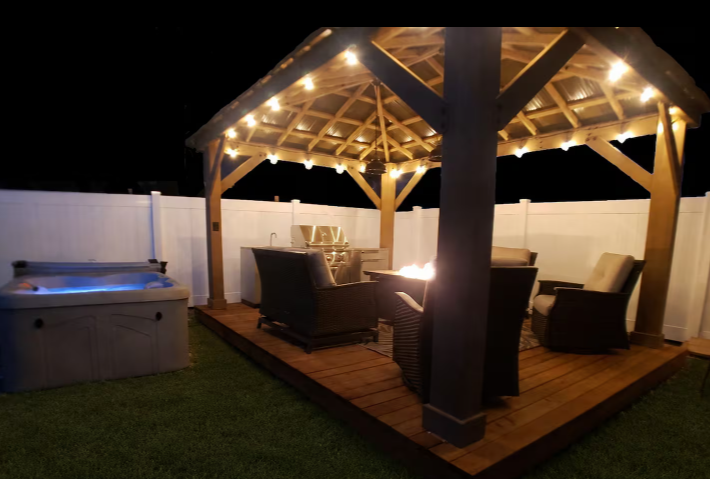 Backyard gazebo with string lights, wicker lounge seating, BBQ grill and private hot tub at night