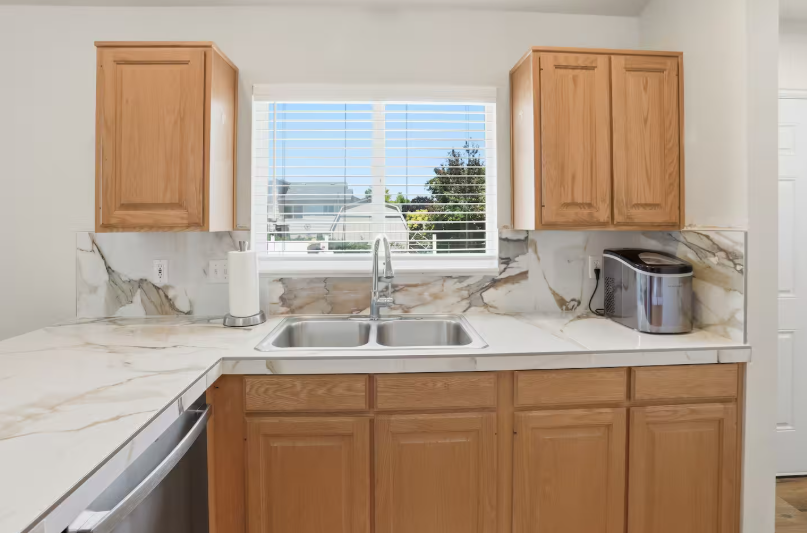 Kitchen sink area with oak cabinets, marble backsplash, double basin sink, ice maker and bright window overlooking backyard