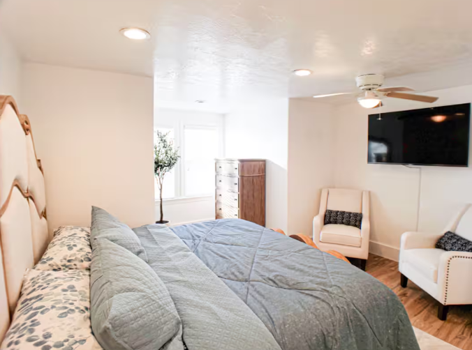 Bright bedroom with upholstered headboard, gray bedding, silver nightstand, and plant
